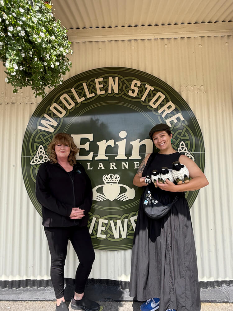 Blarney and Haley Rhodesin standing in front of a sign for 'Woollen Store Erin Larnes', holding toys.